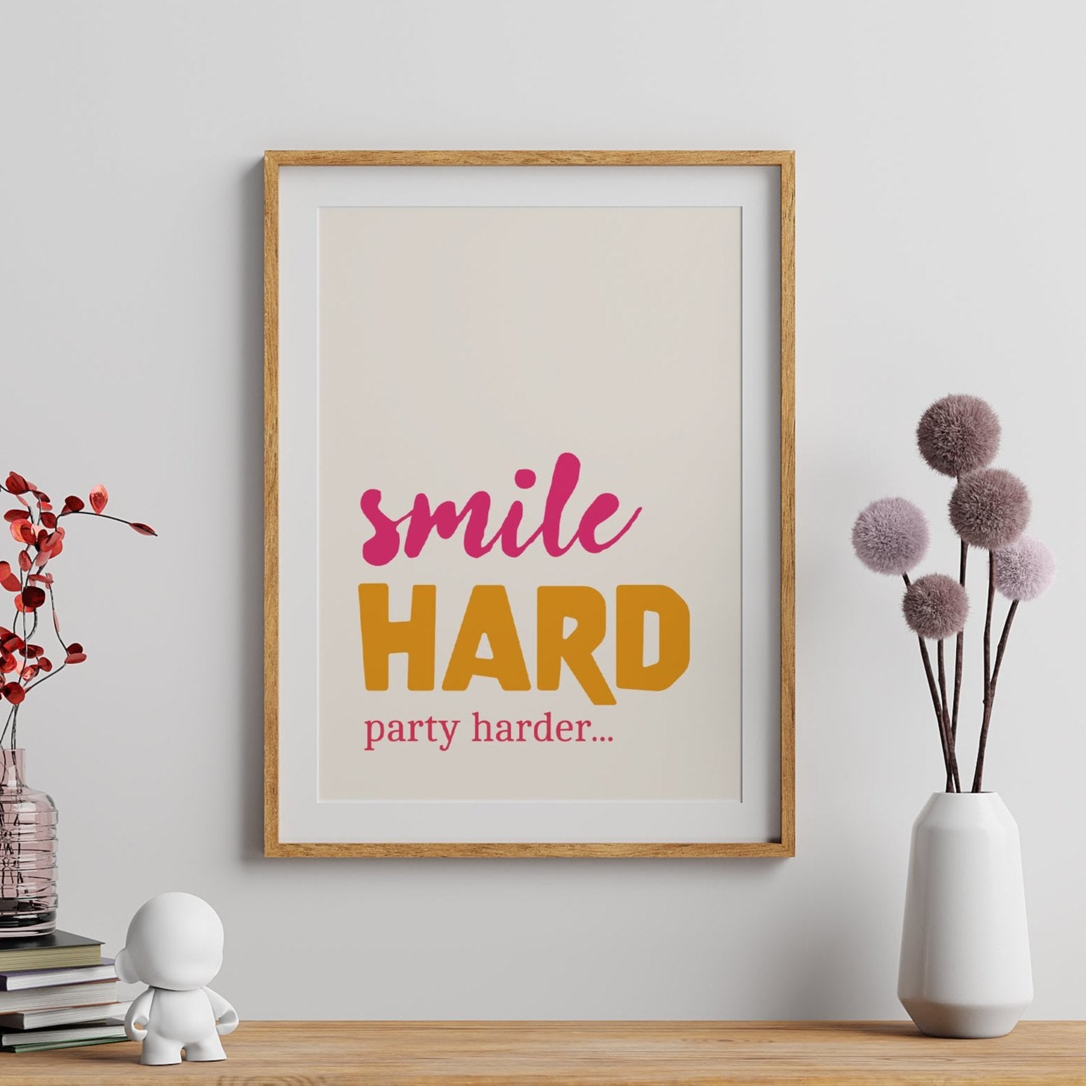 A framed poster with the text 'SMILE HARD PARTY HARDER' in bright pink, orange, and yellow colors, displayed on a wall above a shelf with a few decorative items.