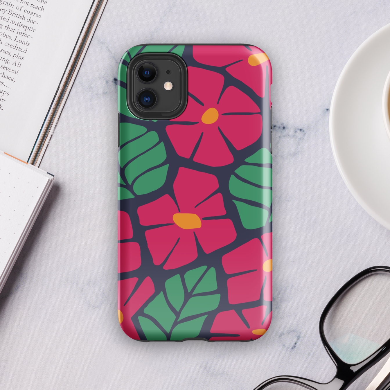 Phone case with colorful floral design on a marble surface with a book, pen, and coffee.